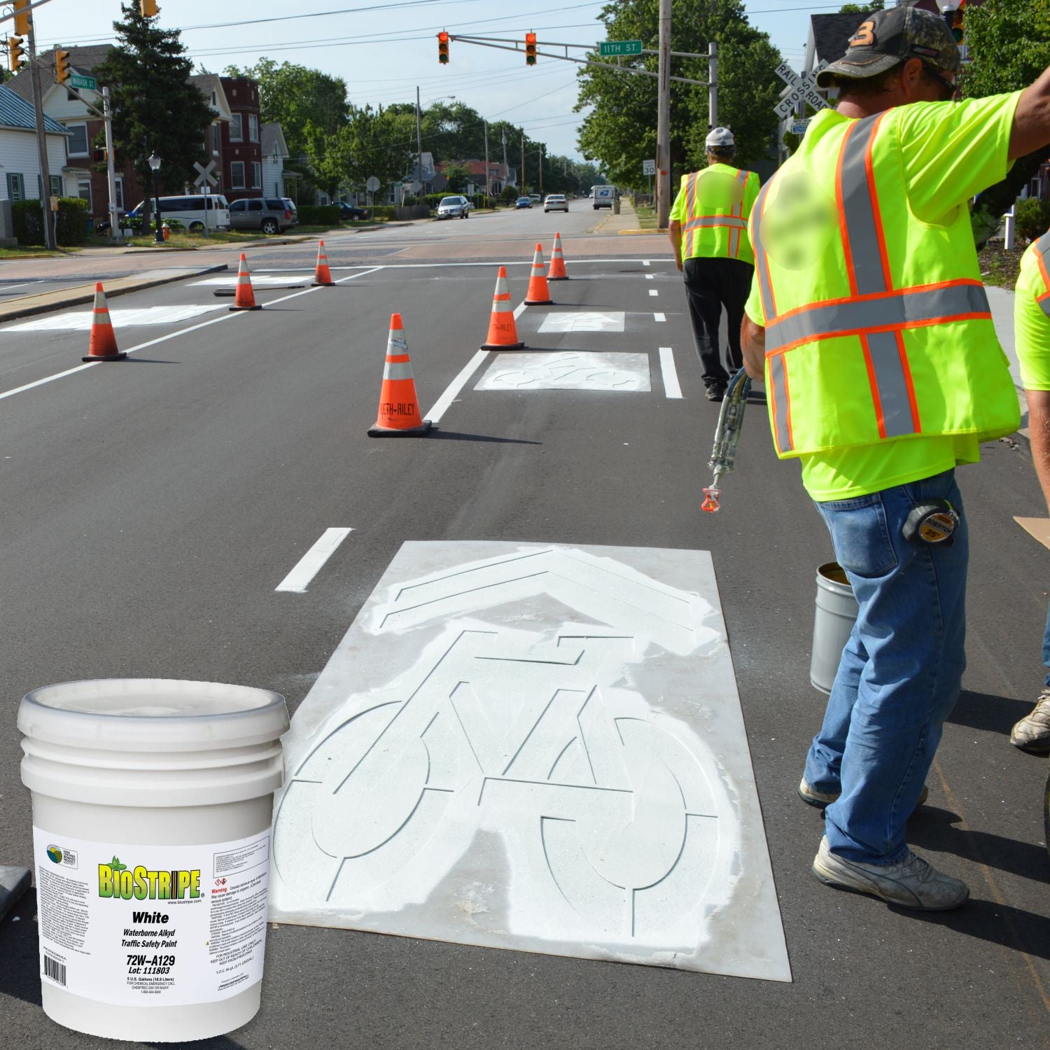 Road crew applying a white bicycle lane stencil on freshly paved asphalt using Aexcel Biostripe Traffic Marking Paint, with traffic cones and workers in high-visibility safety gear.