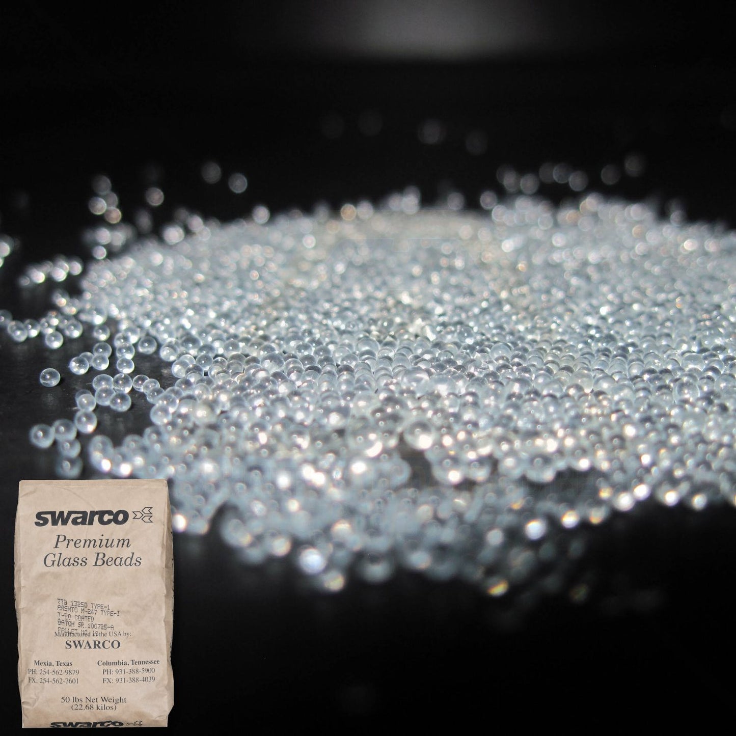 Close-up of SWARCO Premium Glass Beads showing reflective glass spheres used in traffic paint and pavement markings, with a 50 lb bag displayed in the corner.